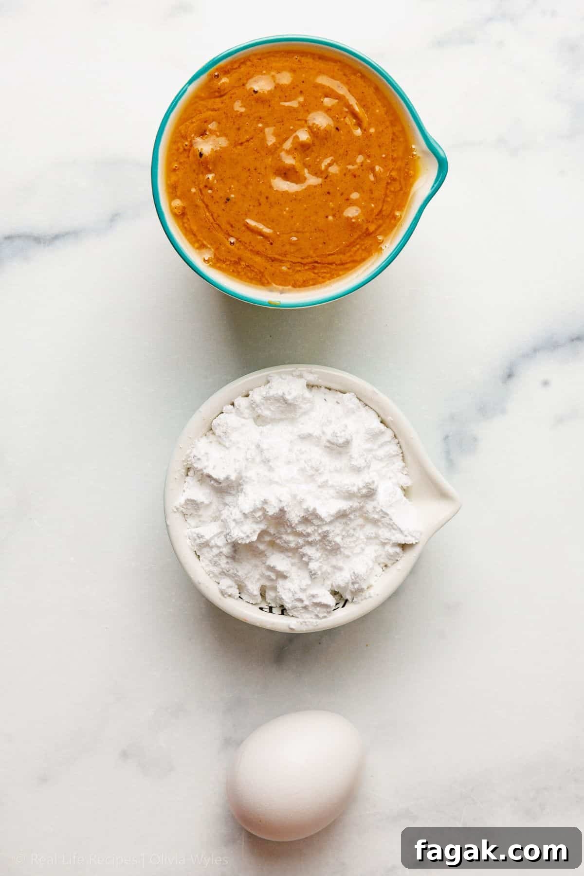 Natural peanut butter in a teal measuring cup, powdered allulose sweetener in a white bowl, and one egg on white marble.