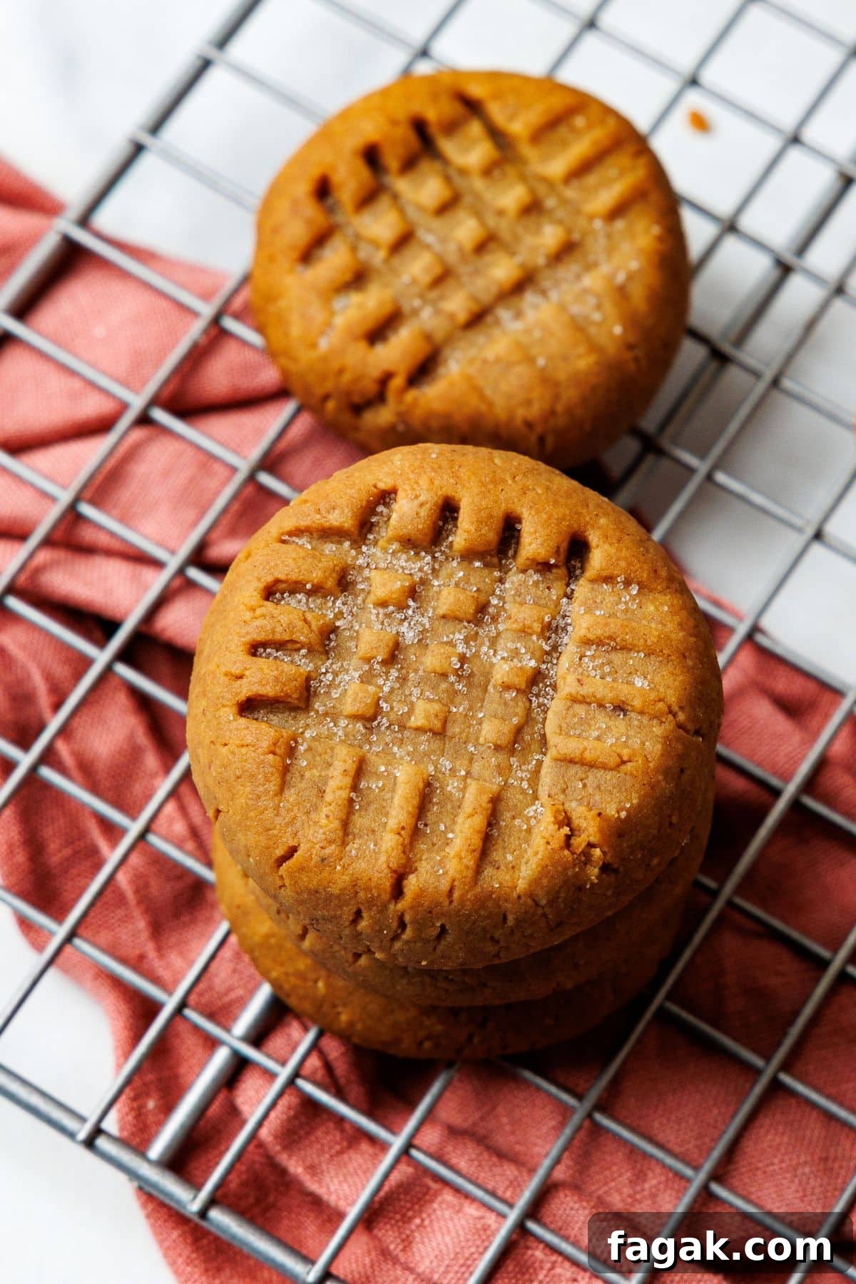 Stack of three sugar-free peanut butter cookies on a wire rack with rust napkin, criss-cross fork pattern on top.