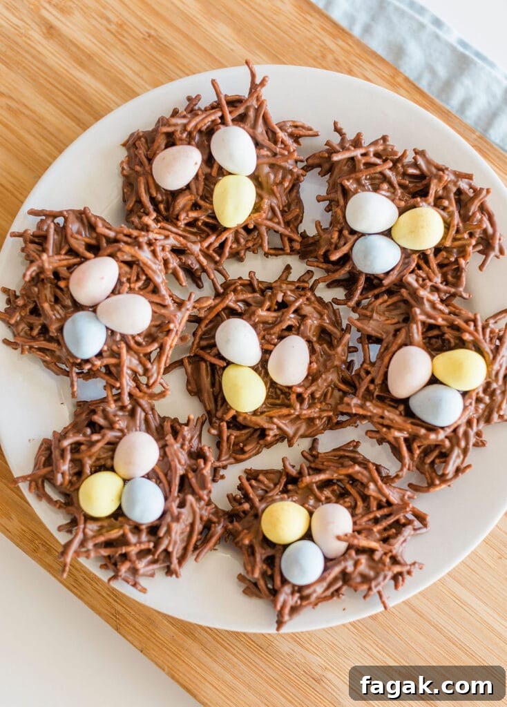 A plate of chocolate nests made from chocolate-coated chow mein noodles, topped with colorful pastel mini eggs, resembling bird nests. The plate is placed on a wooden surface.
