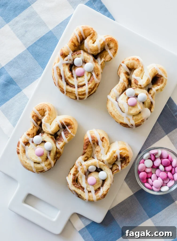 Four bunny-shaped cinnamon rolls with white icing and pink and white candies, arranged on a white cutting board. A small bowl of extra candies is beside them. The board is placed on a blue and white checkered cloth.