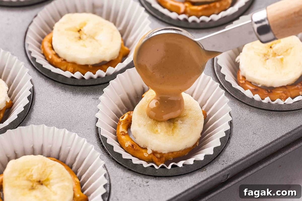 A spoon drizzling peanut butter on a slice of banana butter and pretzel in a muffin case.