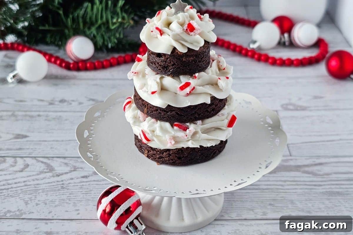 A stack of three round chocolate brownies with white frosting and crushed peppermint, arranged in a tree shape on a white cake stand with holiday decorations in the background.