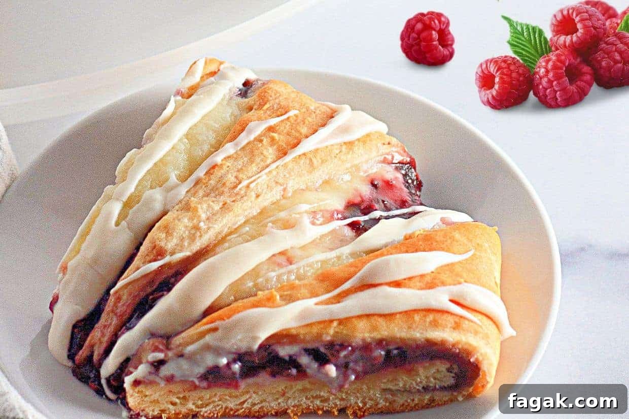 A slice of raspberry crescent ring with white icing on a white plate, with fresh raspberries and a green leaf in the background.