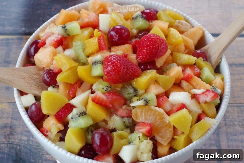 classic fruit salad in a white bowl with wooden serving spoons on each side (on faux wooden surface)