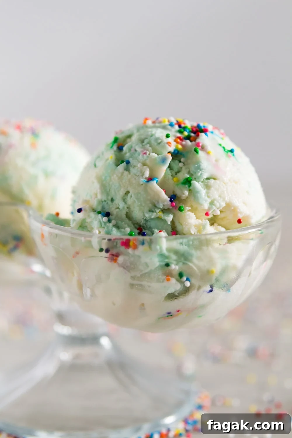 A glass container holding a blue and white scoop of ice cream with sprinkles on top in front of a white background.