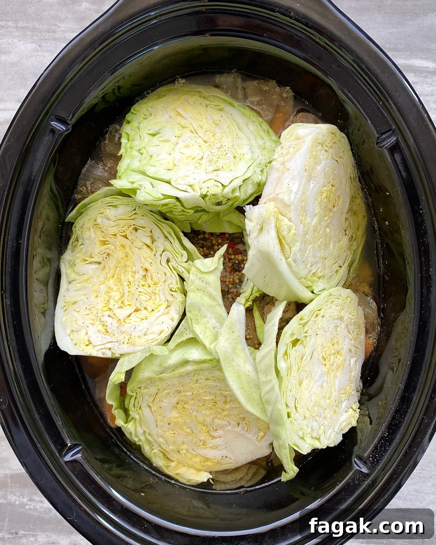 overhead shot of cooked corned beef with potatoes, carrots, and onion in a black slow cooker with uncooked cabbage wedges on top
