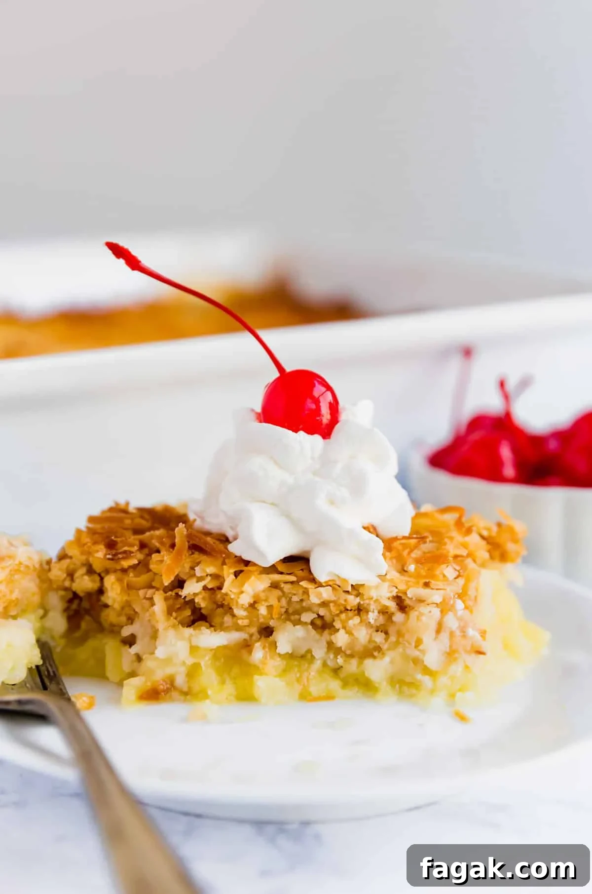 A close up image of Piña Colada dump cake on a plate with whipped cram and a cherry on top on a white plate with a fork.