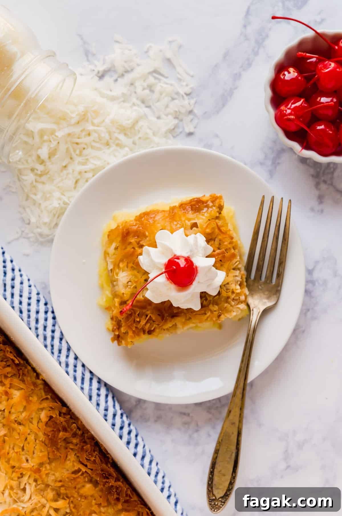 An overhead image of a pice of dump cake on a white plate with a fork on it and coconut and a bowl of cherries next to it.