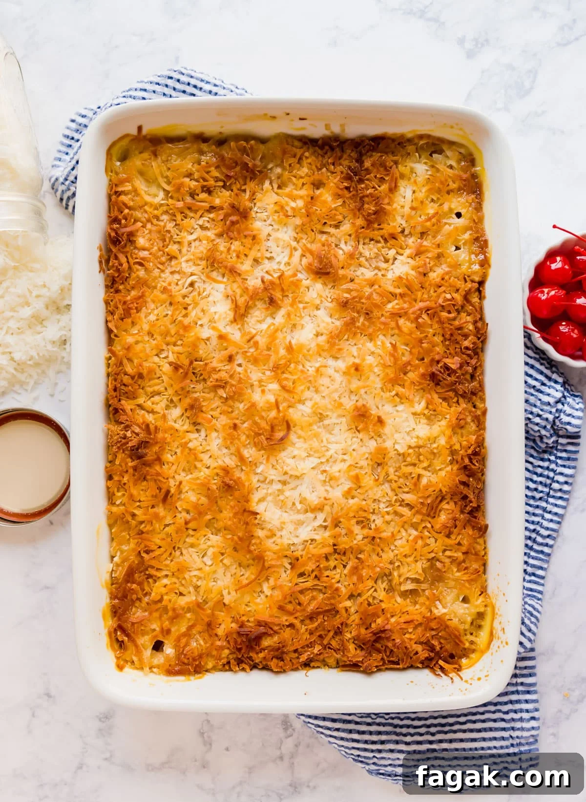 An overhead image of pineapple dump cake in a white baking pan on a blue and white striped towel with coconut flakes and a bowl of maraschino cherries on the sides of it.