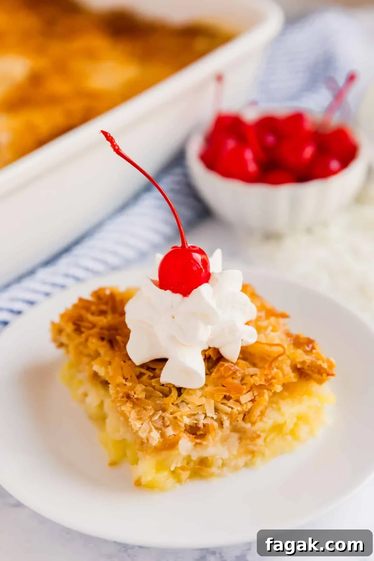 A side angle shot of a pice of dump cake with whipped cream and a cherry on top with a bowl of maraschino cherries and the whole dump cake in the background.