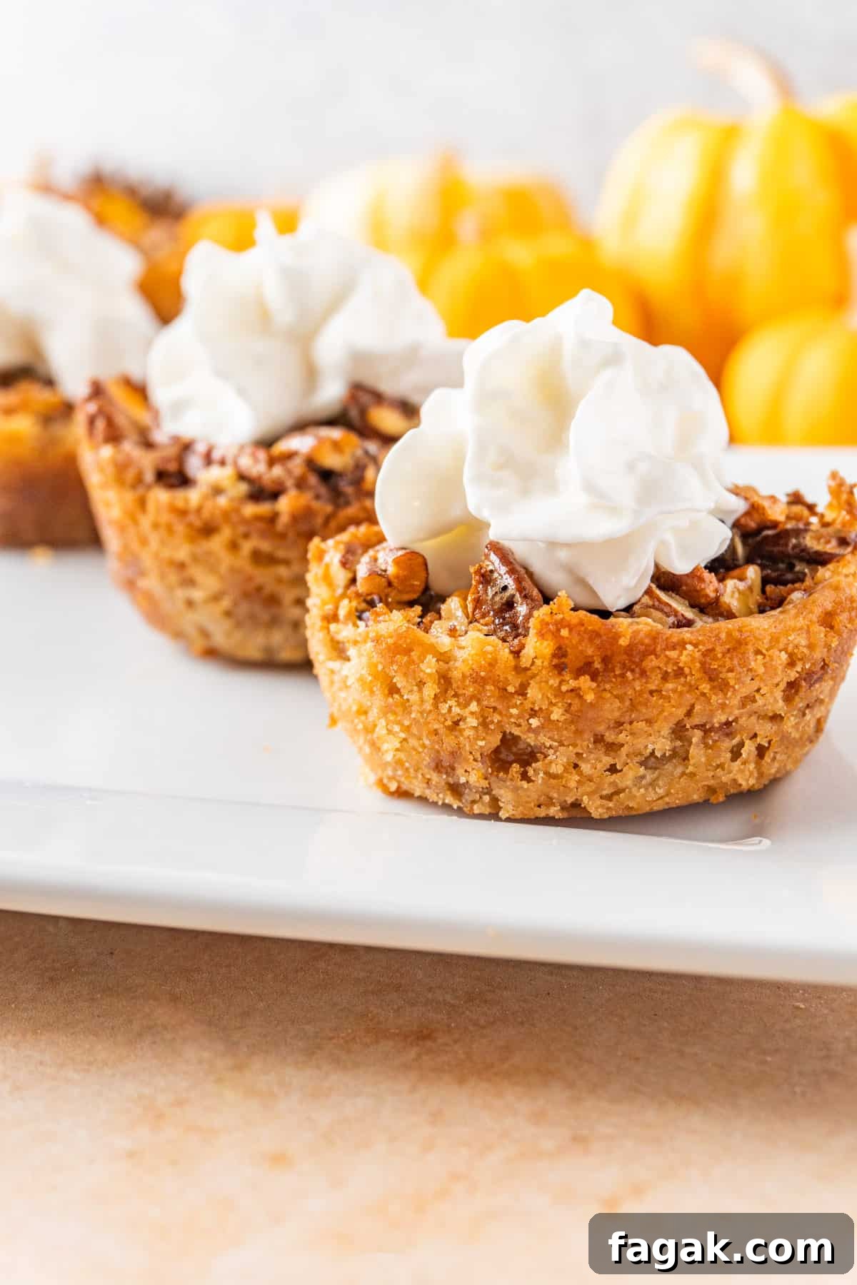 mini pecan pie tarts with whipped cream on top sitting on a white plate with mini pumpkins in the background.