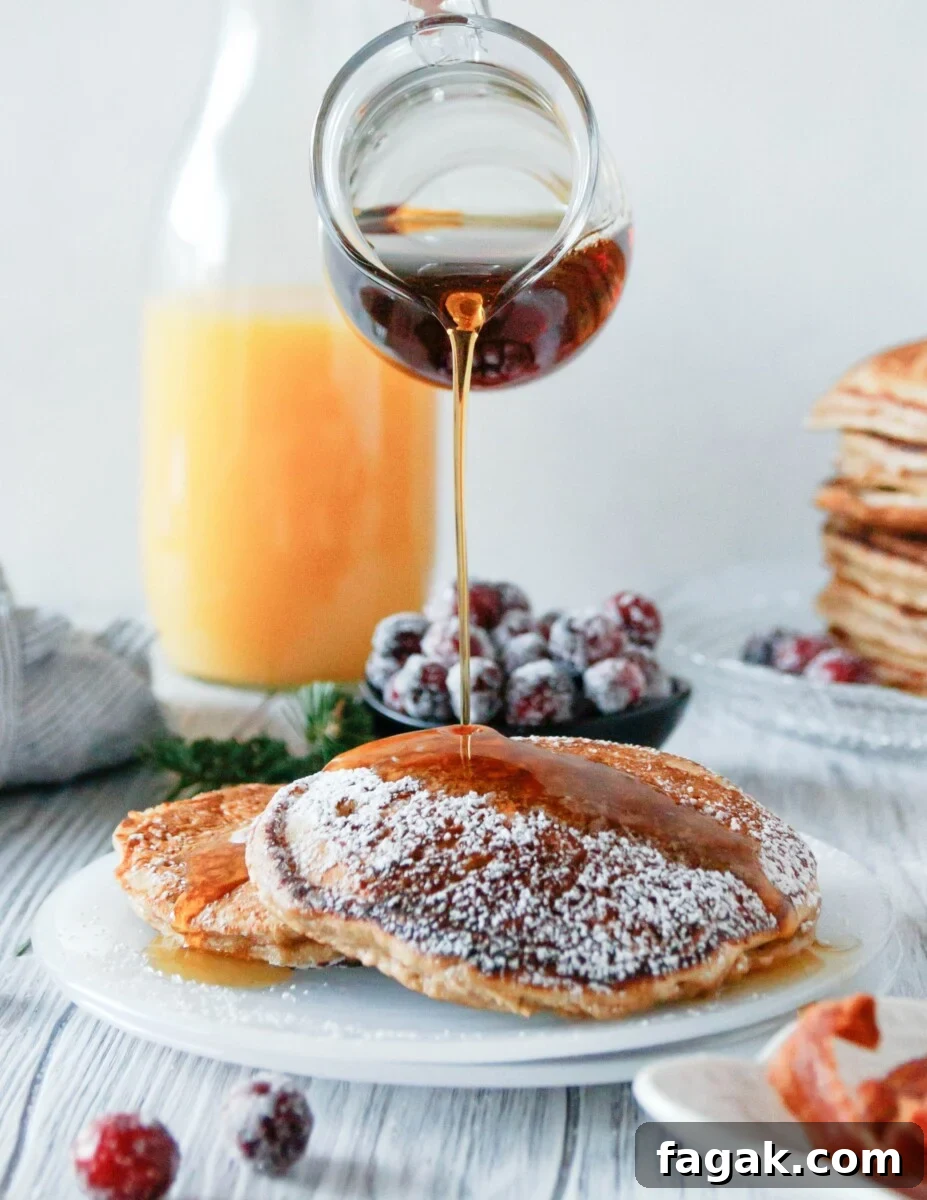 a glass pitcher pouring syrup on plated eggnog pancakes. Orange juice in the background