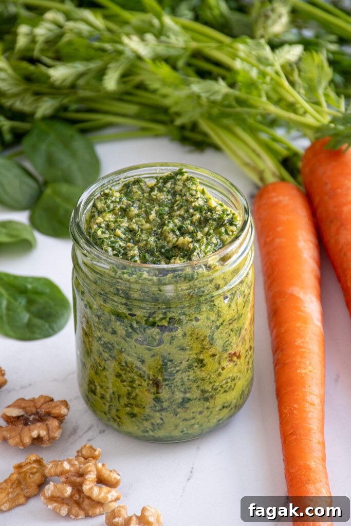 A glass jar filled with green carrot top pesto surrounded by a carrot with greens, spinach, and walnuts.