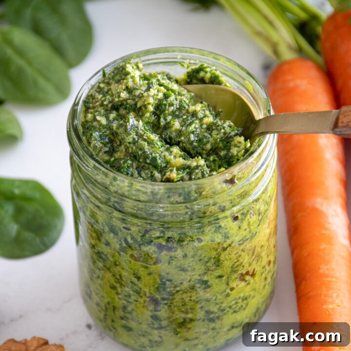 A glass jar with a spoon scooping up green color carrot top pesto.