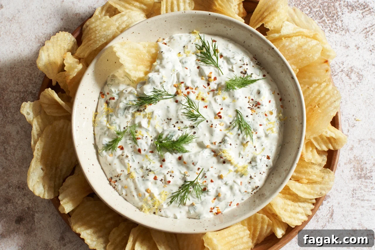 A bowl of dill pickle dip topped with fresh dill, pepper, and lemon zest, surrounded by ridged potato chips.