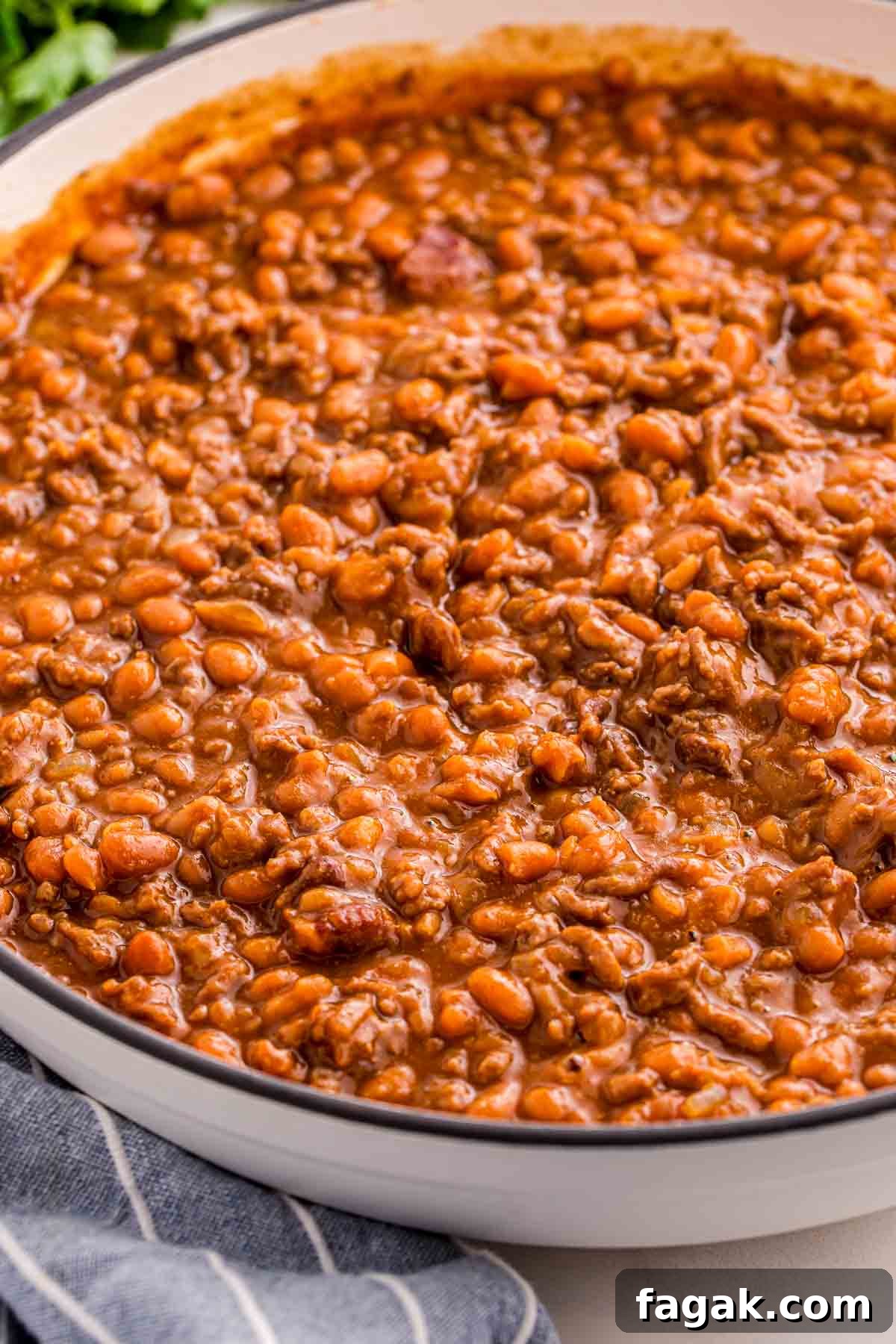 Close up of baked beans with ground beef in a rich reddish brown sauce filling a shallow white bowl. The thick bean mixture appears hearty and glossy with visible crumbles of beef throughout.