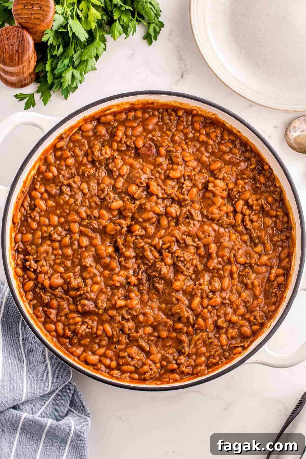 Overhead view of a large pan filled with baked beans and ground beef in a thick savory sauce. Fresh parsley and serving utensils surround the dish on a light countertop.