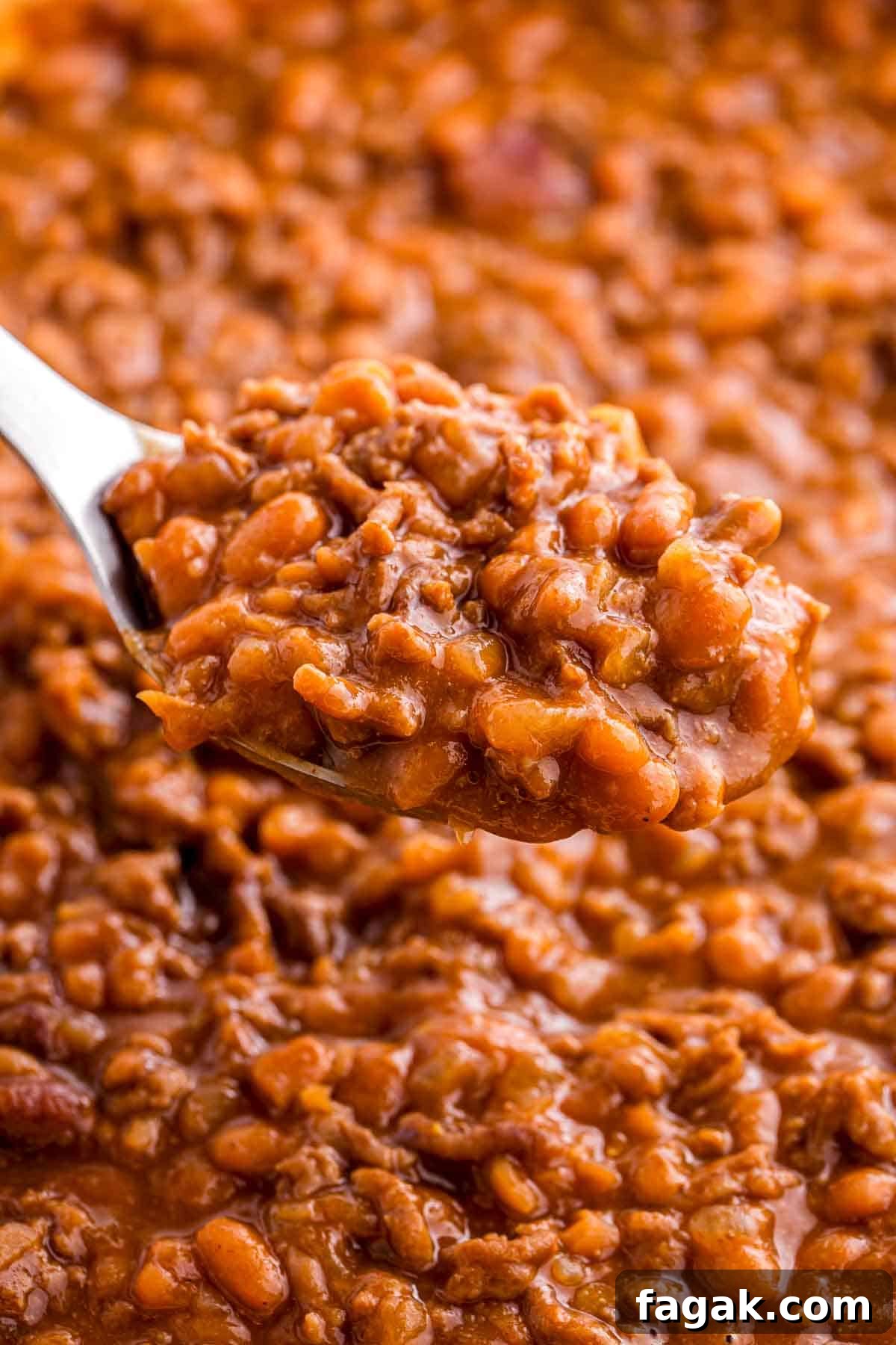 Spoon lifting a thick scoop of baked beans with ground beef from a pot. The close up highlights the saucy texture, tender beans, and crumbled beef.