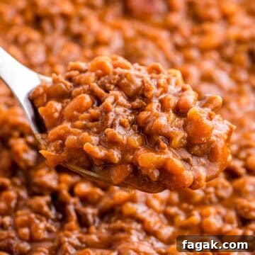 Spoonful of baked beans with ground beef held above the dish to show the thick texture and rich sauce. Tender beans and crumbled beef are visible in the close up.