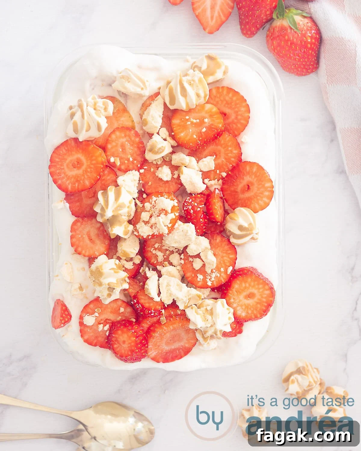A photo from above of a bowl of strawberry tiramisu topped with strawberry slices and crumbled meringue. At the bottom two golden spoons and meringue. Extra strawberries on top. A white background.