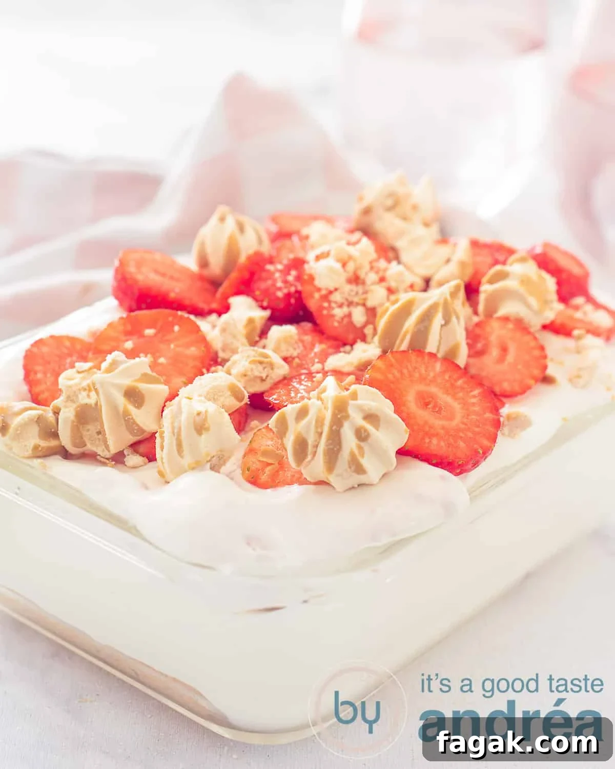 An elevation view of a glass bowl filled with strawberry tiramisu with a strawberry and meringue garnish. a white background and a pink and white tea towel in the background