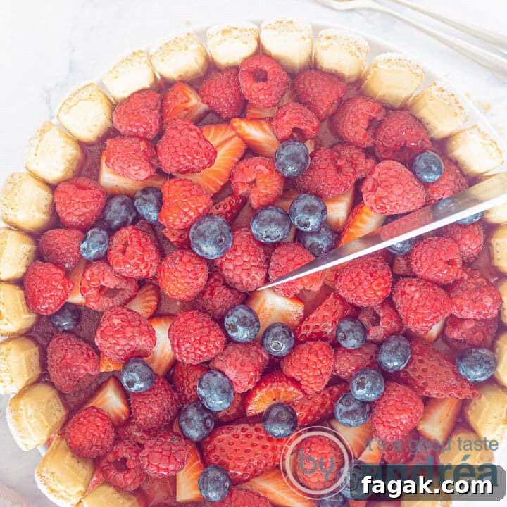 A square photo of a charlotte russe from above. A topping of raspberries, blueberries and strawberries. A knife cuts off a wedge.