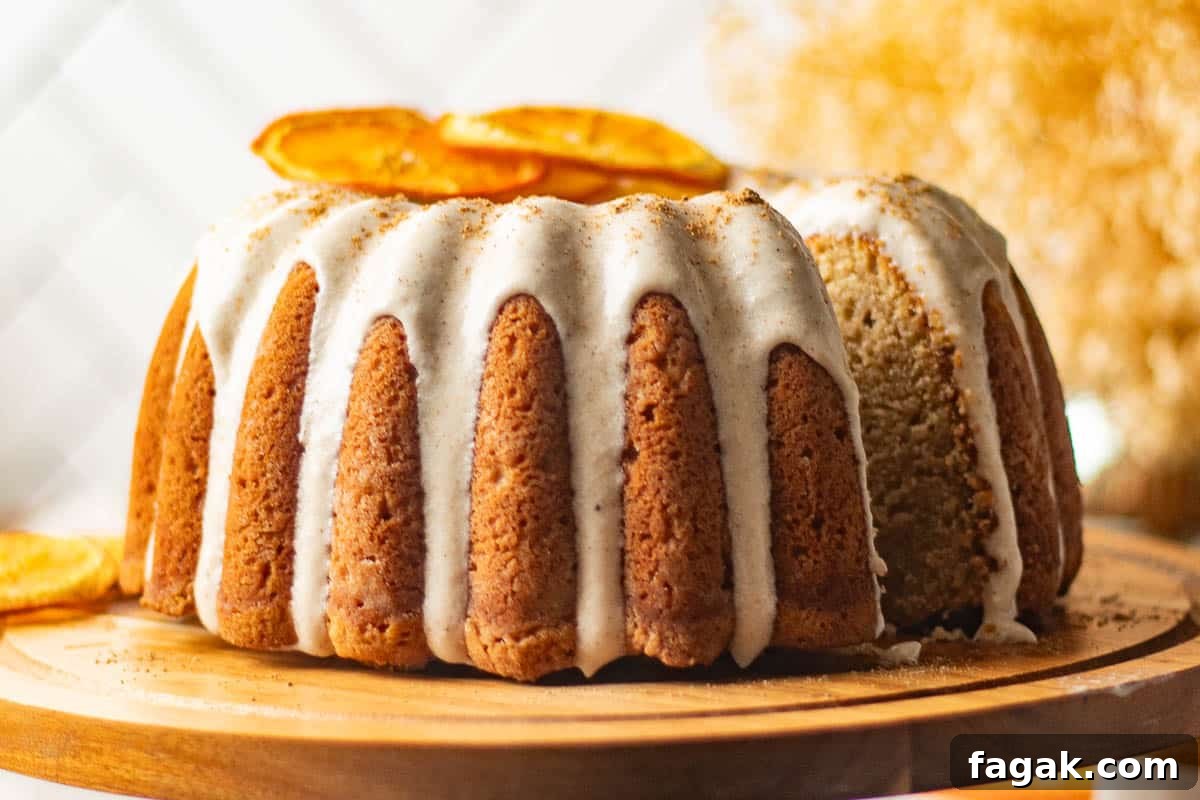 A chai bundt spice cake on a wooden stand, featuring a thick layer of cream cheese frosting and dried orange slices as a garnish, with a cozy, warm backdrop.