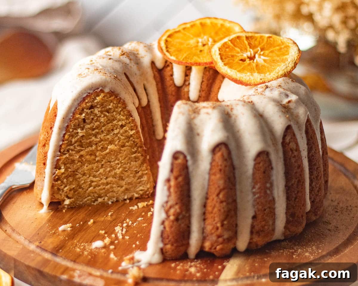 A beautifully presented chai bundt spice cake on a wooden stand, with a slice removed to reveal the soft, spiced crumb inside, highlighting the cream cheese icing and orange garnish.