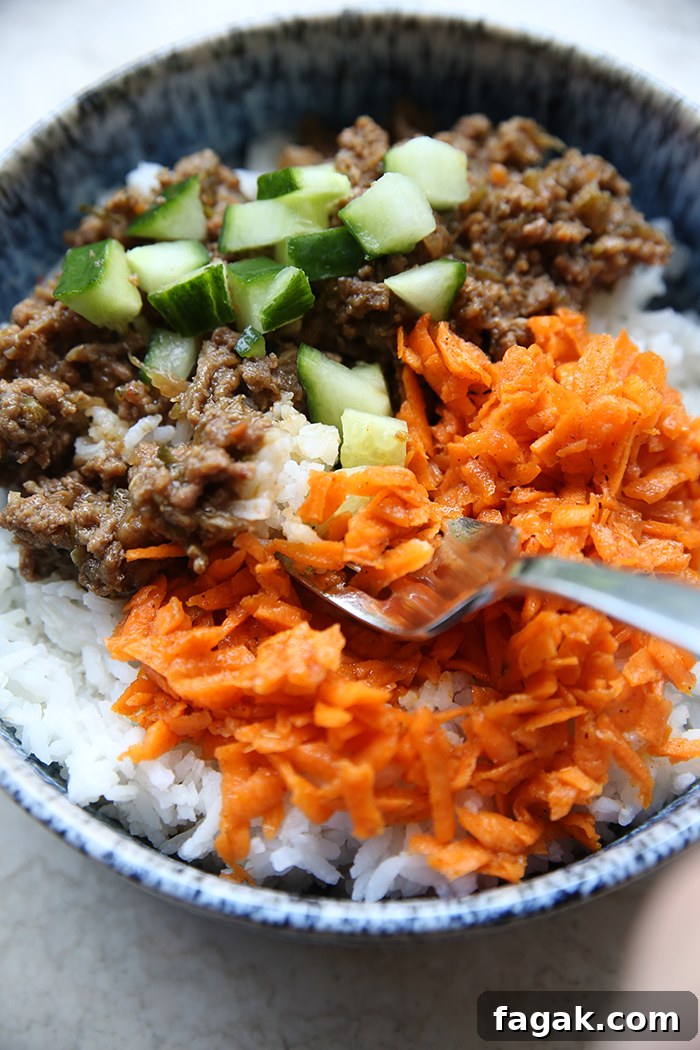 A bowl of rice with chopped cucumbers on top of ground beef and next to carrot kimchi. A spoon is inserted diagonally from the right into the carrot kimchi.