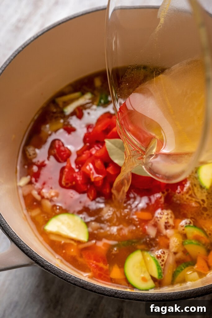 Vegetable broth poured over minestrone soup ingredients in a large pot.