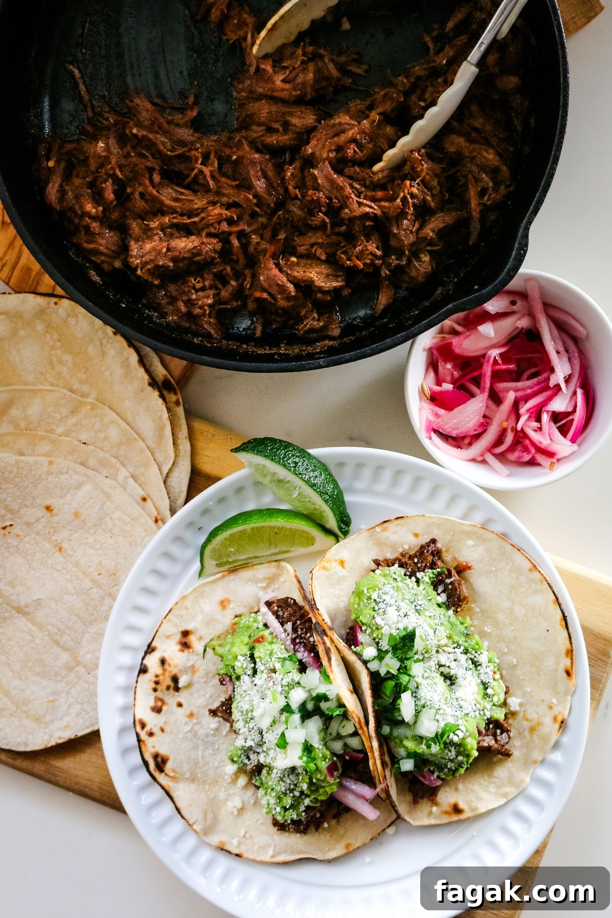 Slow-Braised Chuck Roast Tacos with Salsa Verde 2 Roast tacos on a white plate with roasted beef in a pan, with tacos and onions beside it.