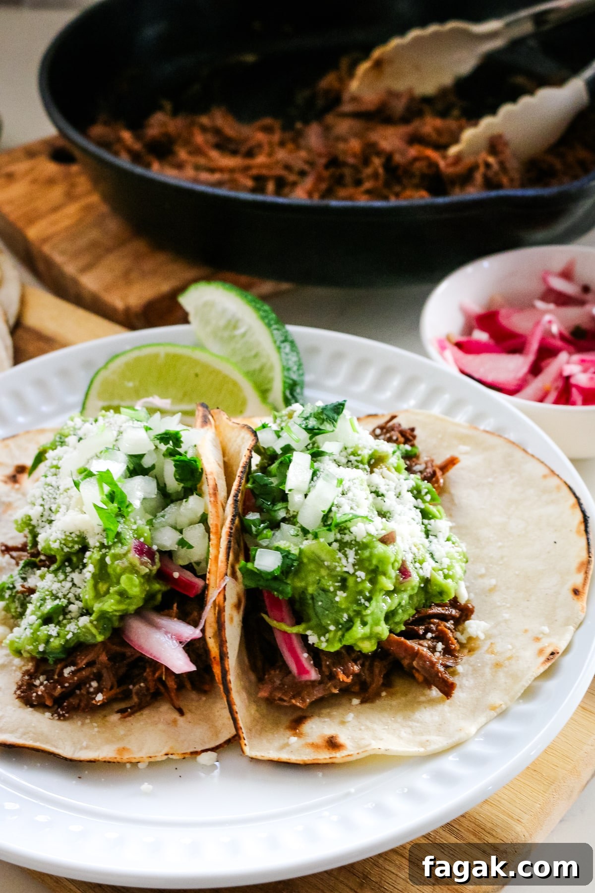 Slow-Braised Chuck Roast Tacos with Salsa Verde 5 Roast tacos on a white plate with roasted beef in a pan beside them.