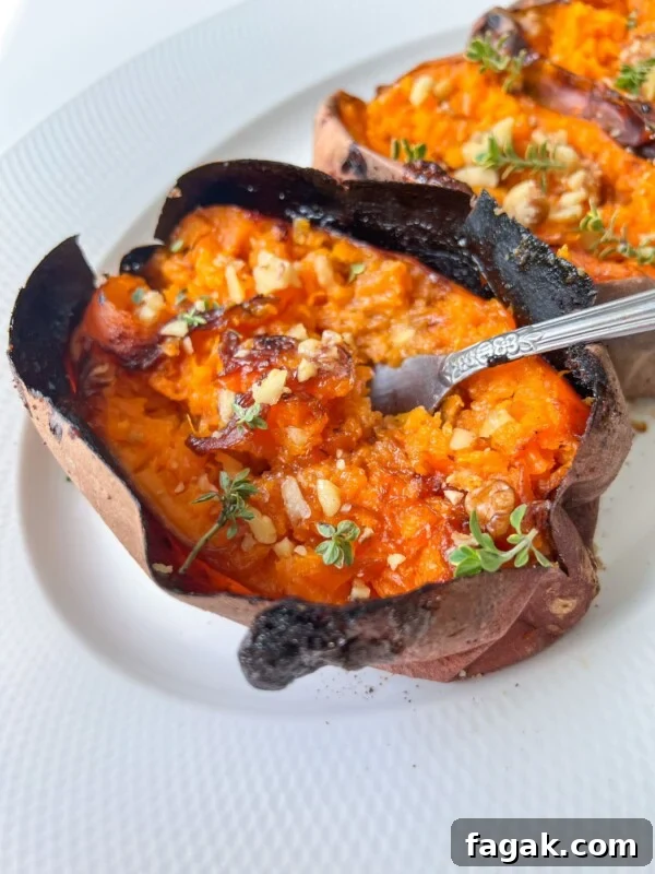 A baked sweet potato split open and topped with herbs and chopped nuts, served on a white plate. A fork is inserted into the soft, orange flesh of these crispy roasted sweet potatoes.