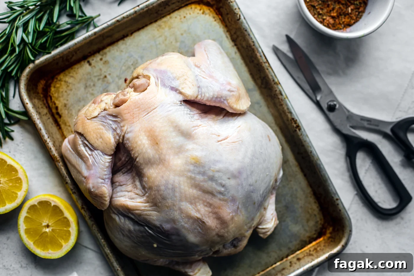 Baking sheet with raw whole chicken next to kitchen shears, seasoning, lemon halves, and fresh herbs.