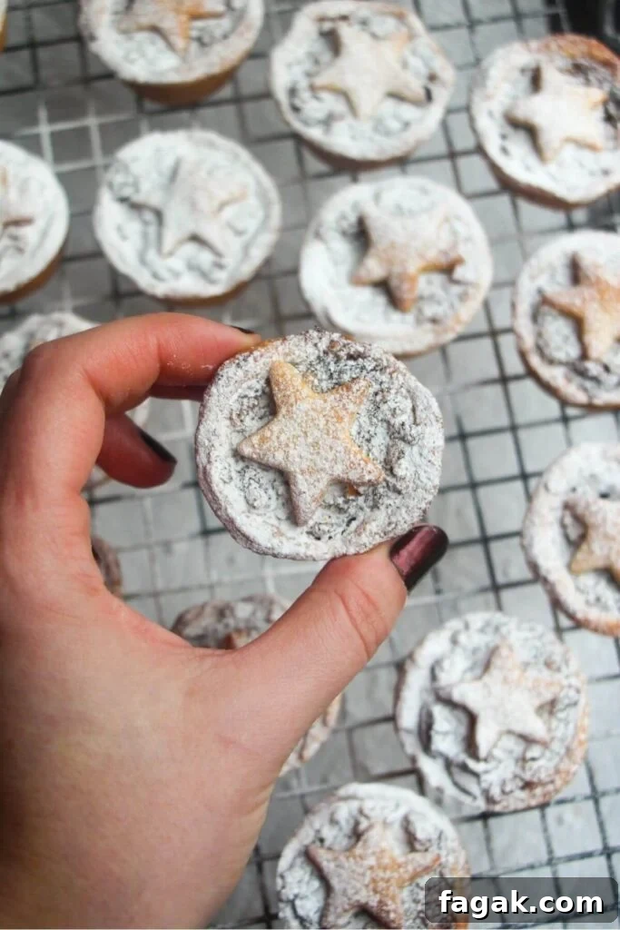 A hand holding a Christmas fruit mince pie dusted with icing sugar with more mince pies in the background.