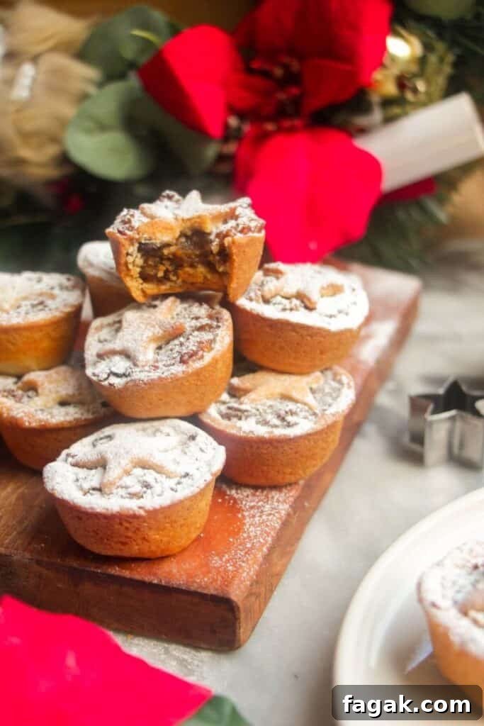 A pile of icing sugar dusted Christmas mince pies on a small wooden board with Christmas deocrations in the background.