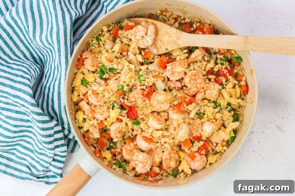 16 Light and Satisfying Shrimp Recipes for Quick Weeknight Meals 17 A skillet brimming with keto fried cauliflower rice, shrimp, peas, carrots, and bell peppers is accompanied by scrambled eggs. A wooden spoon rests on the side, while a striped cloth adds charm next to the skillet.