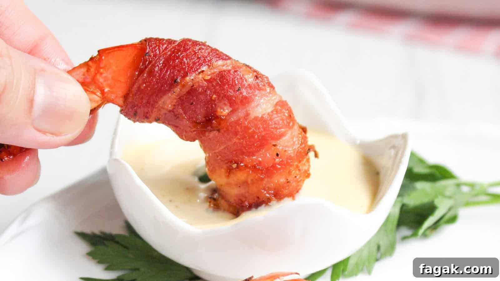 16 Light and Satisfying Shrimp Recipes for Quick Weeknight Meals 5 A hand holding a bacon-wrapped shrimp dipped in a small bowl of sauce, with a green leaf garnish on the side.