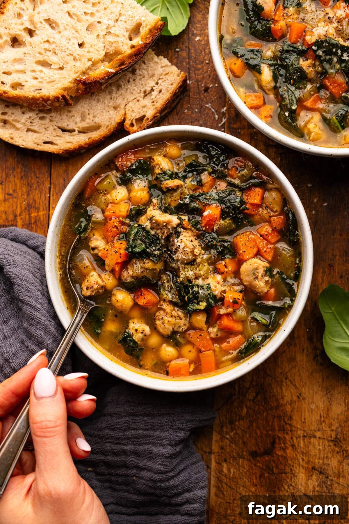 A hand holding a spoon in a bowl of Italian sausage soup. The bowl of soup is arranged on a wooden board with slices of bread and a dark gray napkin around it.
