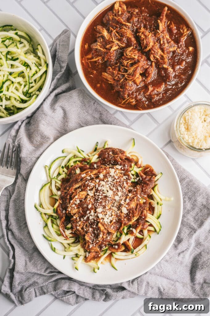 Overhead photo of balsamic chicken with zoodles on a white plate. At the top of the photo are two bowls, one filled with zoodles, the other filled with balsamic chicken. There is a small glass jar filled with parmesan cheese, too.