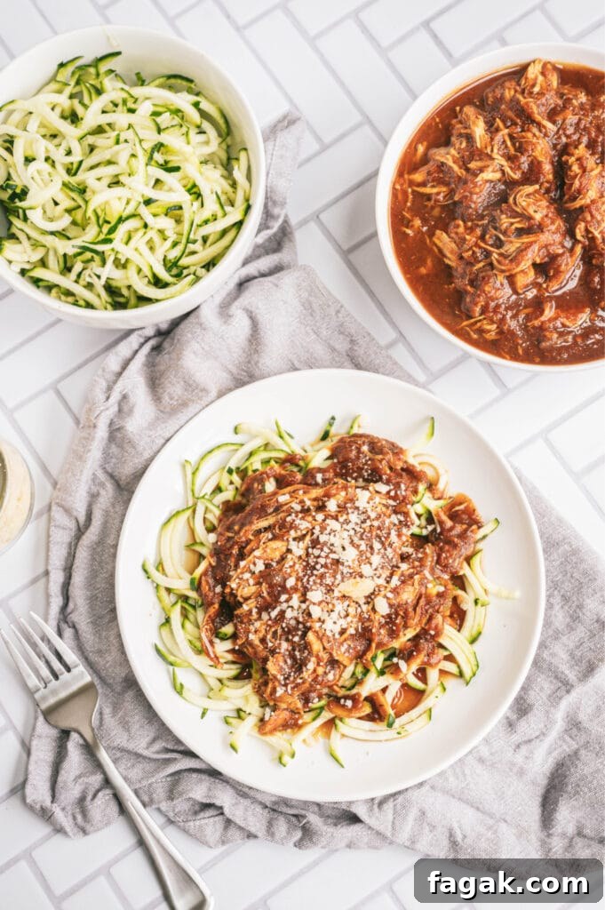 Close up overhead photo of chicken with zoodles on a white plate on a gray kitchen towel. There is a bowl of zoodles and a bowl of chicken on the side.