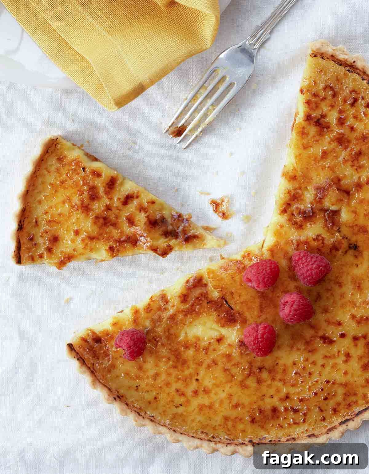 A partially sliced coconut crème brûlée tart topped with raspberries on a white linen cloth with a fork and napkin beside it.
