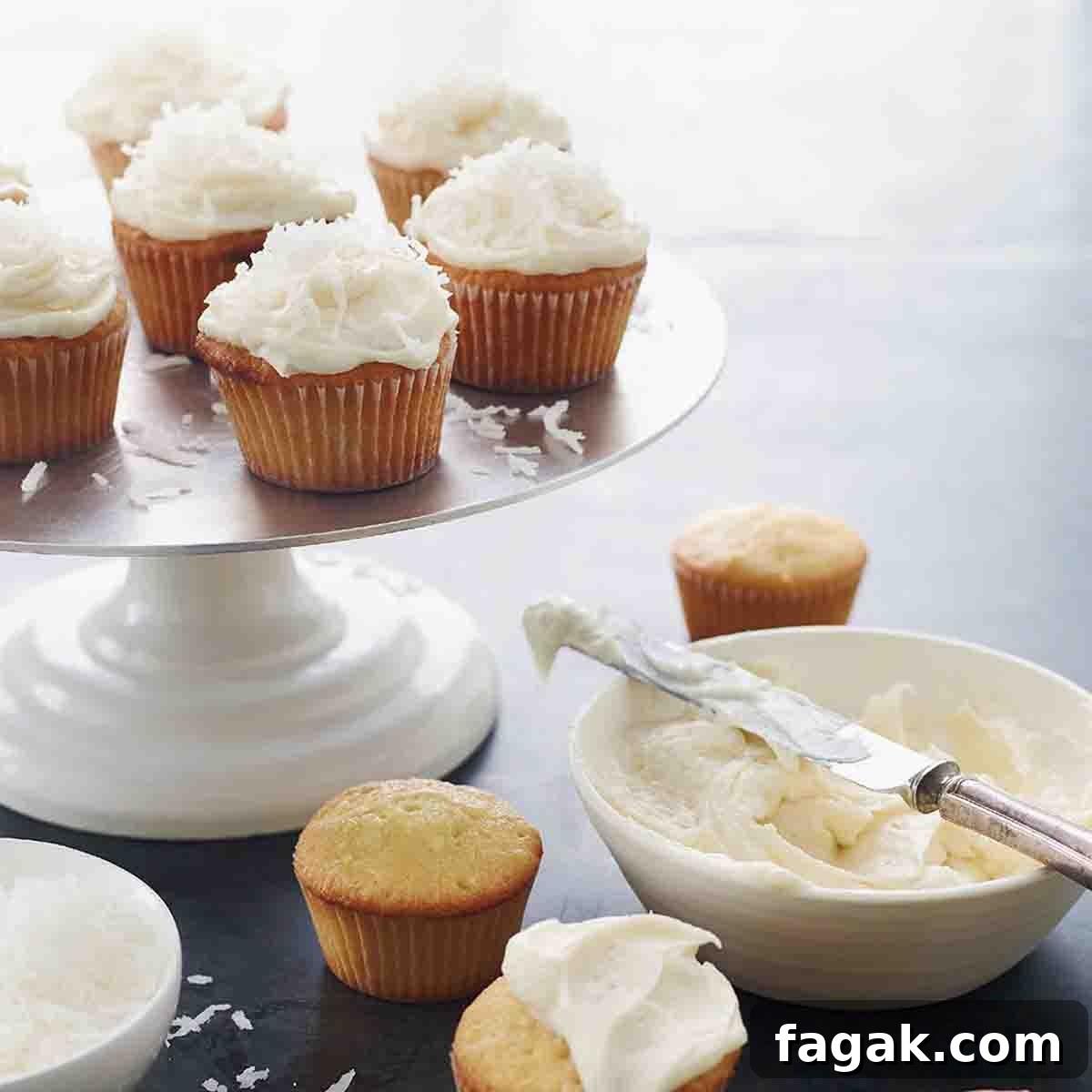 Cake stand with 7 frosted coconut cupcakes, four unfrosted cupcakes on the table, and a bowl of frosting.