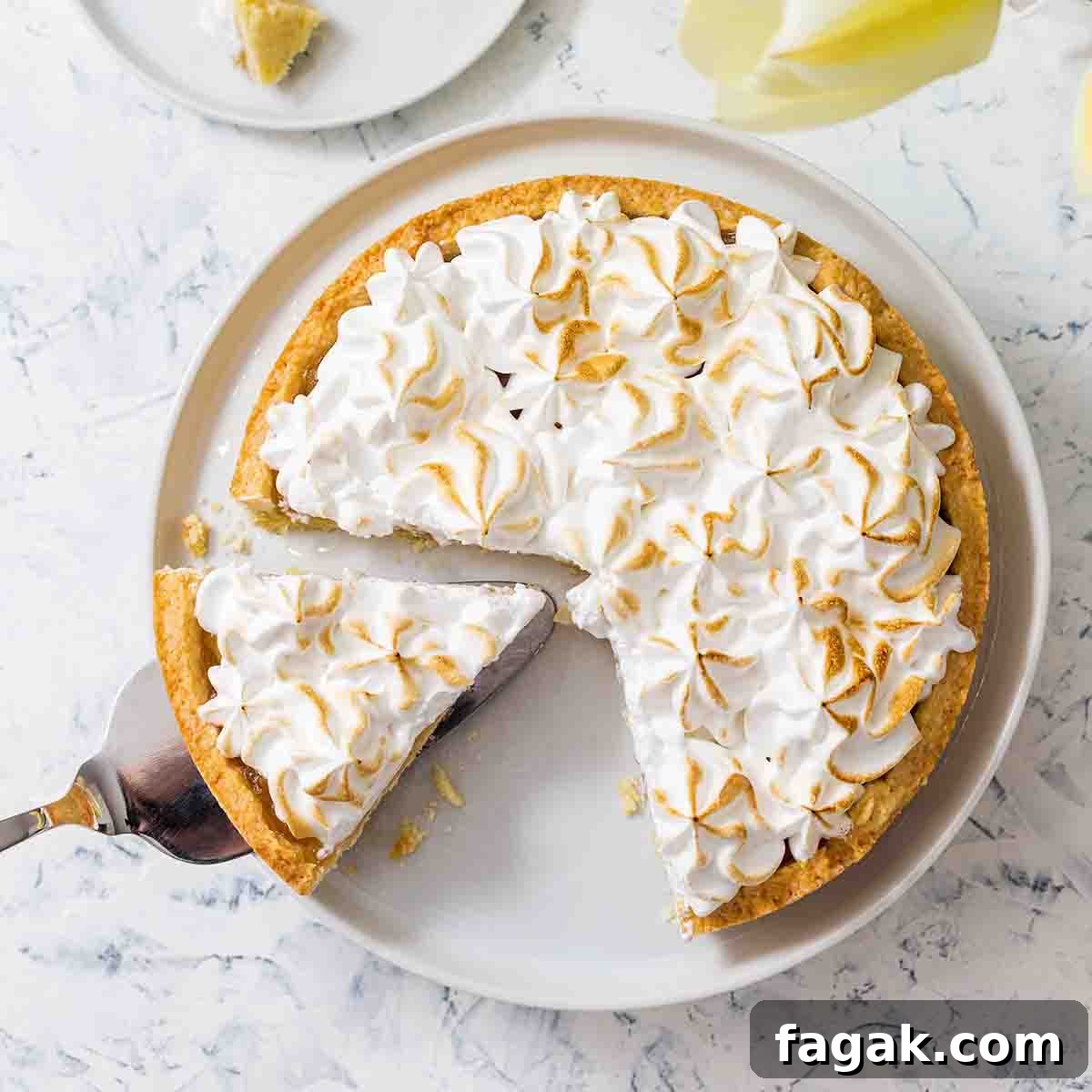 A lemon meringue tart in a white dish, with one slice on a plate beside it and one more being lifted out.