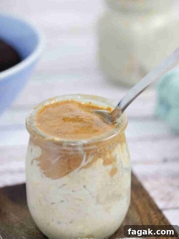 Mason jar with espresso overnight oats topped with almond butter and a spoon in it on a wooden coaster with another jar of oats and a bowl of coffee behind it.