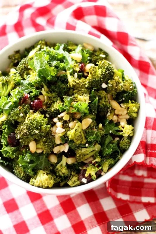 Wooden table with a white bowl sitting on it containing a kale and broccoli salad, red and white gingham napkin on table.