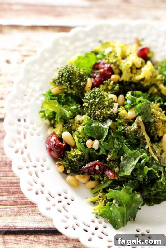 White plate sitting on a wooden table containing a kale and broccoli salad with pine nuts and dried cranberries.
