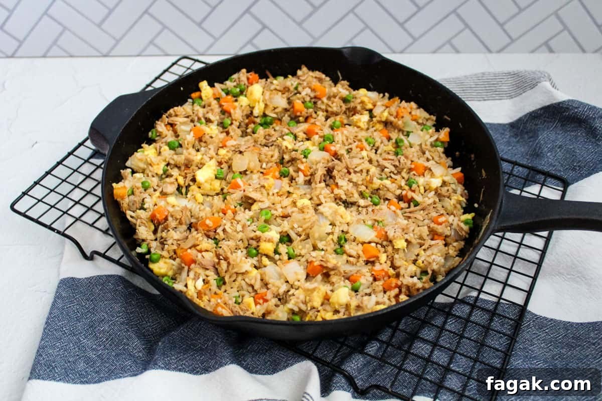 A cast iron skillet filled with fried rice containing eggs, peas, carrots, onions, and pulled pork on a wire rack with a kitchen towel under it.