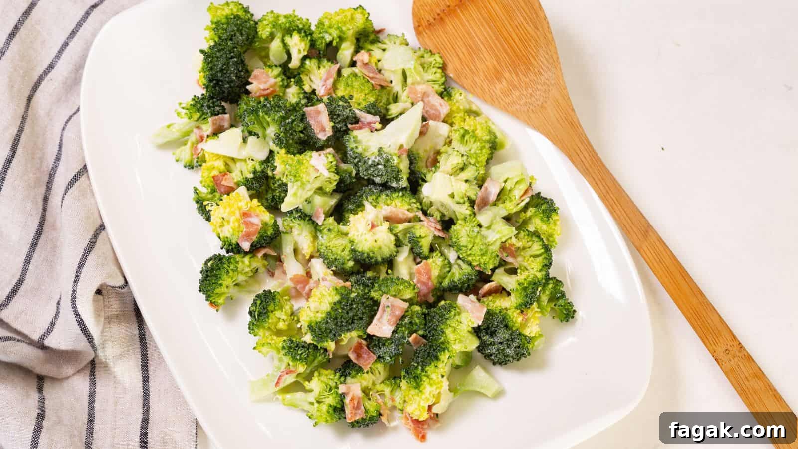 Broccoli salad with bacon bits on a white oval plate, accompanied by a wooden serving spoon on a linen napkin.