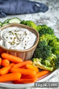 Ranch dip in a gray bowl surrounded by veggies on a white counter with blue towel in background.
